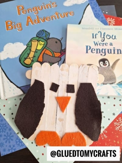 A handmade popsicle stick penguin, crafted from felt and paper, sits atop two children’s penguin books—Penguins Big Adventure and If You Were a Penguin—with colorful paper beneath.