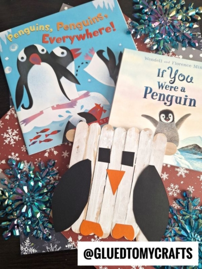 Two children’s books about penguins, sparkly blue snowflake decorations, and a fun craft stick penguin made from popsicle sticks, construction paper, and glue are displayed together. @GLUEDTOMYCRAFTS text is at the bottom.