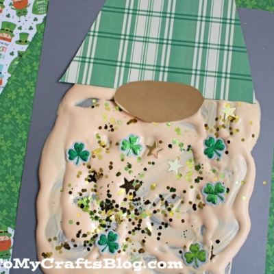 A puffy paint leprechaun craft features a plaid green paper hat, beige oval face, and a fluffy puffy paint beard with shamrock shapes and glitter, all displayed on overlapping colorful paper sheets.