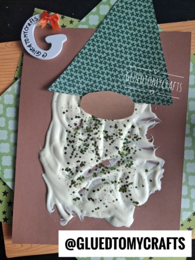 A puffy paint leprechaun craft made with a green patterned hat, oval nose, and a beard created from white glue and glitter on brown paper. A G ornament and patterned paper are in the background. @GLUEDTOMYCRAFTS is shown.