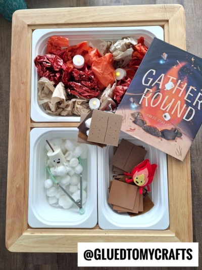 A wooden tray holds three bins: one with paper flames and LED tea lights, one with craft supplies like cotton balls and a marshmallow on a stick for pretend play smores, and one with paper squares. A book titled Gather Round is nearby. @gluedtomycrafts.