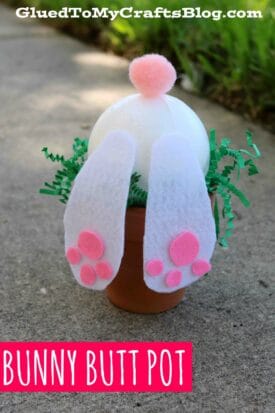 A small terra cotta pot filled with green shredded paper holds a white ball topped with a pink pom-pom and decorated with felt bunny feet and pink toe pads, creating a cute Terra Cotta Pot Bunny Butt. Text reads Bunny Butt Pot.