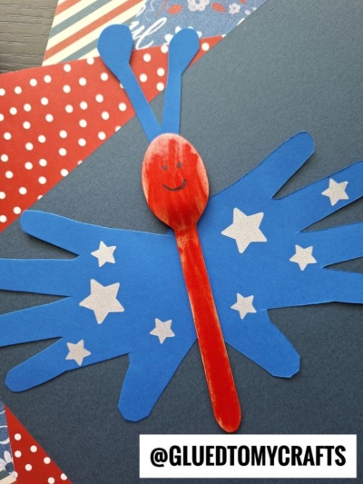 A red wooden spoon with a smiling face is decorated as a handprint butterfly puppet, featuring blue paper wings with white star stickers, set against red polka dot and blue paper. @GLUEDTOMYCRAFTS watermark appears at the bottom.