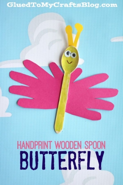 A butterfly craft made from a yellow wooden spoon for the body and pink paper handprints for wings, with googly eyes and antennae, on a blue background with clouds. This adorable handprint butterfly puppet is perfect for creative kids’ projects. Text: HANDPRINT WOODEN SPOON BUTTERFLY.