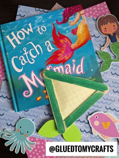 A children's book titled How to Catch a Mermaid sits on a table surrounded by paper cutouts of a mermaid, fish, octopus, popsicle stick mermaid tails, and a green and yellow mermaid tail craft. Handle @GLUEDTOMYCRAFTS is at the bottom.
