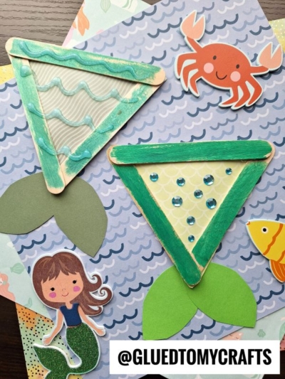 Two popsicle stick mermaid tails crafted with patterned paper rest on ocean-themed paper. Decorative cutouts of a mermaid, crab, and fish surround the crafts. Text reads @GLUEDTOMYCRAFTS.