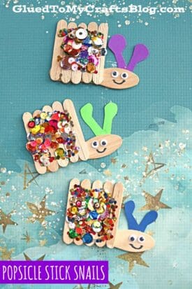 Three snail kid crafts made from popsicle sticks, sequins, and colored paper are displayed on a blue background with gold stars. Each craft stick snail has a smiling face, colorful antennae, and a label reading “Popsicle Stick Snails.”.