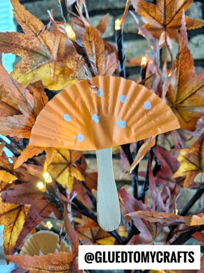 An orange cupcake liner mushroom with white dots and a craft stick forms a cute mushroom craft for kids, hanging among artificial autumn leaves and string lights. Text at the bottom reads @GLUEDTOMYCRAFTS.