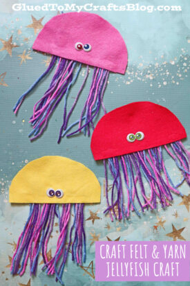 Three colorful Felt Jellyfish crafts made from felt and yarn, each with googly eyes, are arranged on a blue star-patterned background. The yarn jellyfish come in pink, red, and yellow with multicolored yarn tentacles.