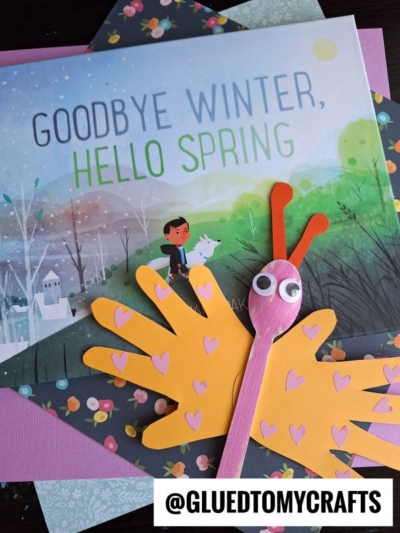 A children’s book titled Goodbye Winter, Hello Spring is surrounded by pastel craft papers. In front is a handprint butterfly puppet made from a pink spoon, yellow paper hands for wings, and pink heart stickers.