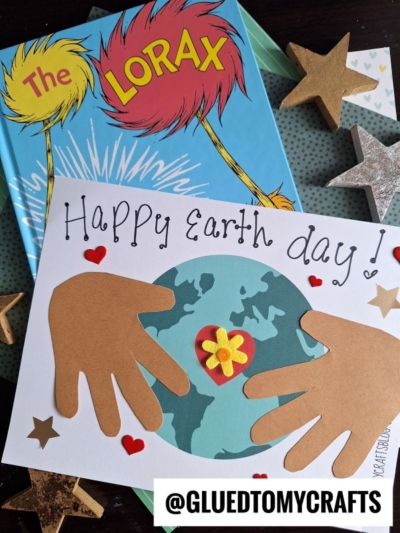 A handmade handprint Earth Day card with paper hand cutouts holding a globe, surrounded by hearts and stars, placed on The Lorax book with wooden star decorations. @GLUEDTOMYCRAFTS is written at the bottom.