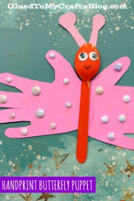 A handprint butterfly puppet made using a painted wooden spoon for the body and pink paper handprint wings, decorated with shiny sequins. Set on a blue starry background. Text reads “HANDPRINT BUTTERFLY PUPPET.” A fun spoon butterfly puppet craft!.