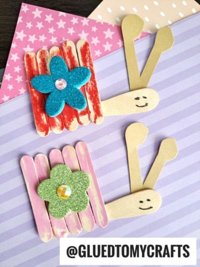 Two popsicle stick snails made from colored sticks and foam flowers sit on pastel striped and polka dot paper. Each snail has a smiling face drawn on its head. The text @GLUEDTOMYCRAFTS appears at the bottom, highlighting this fun snail kid craft.