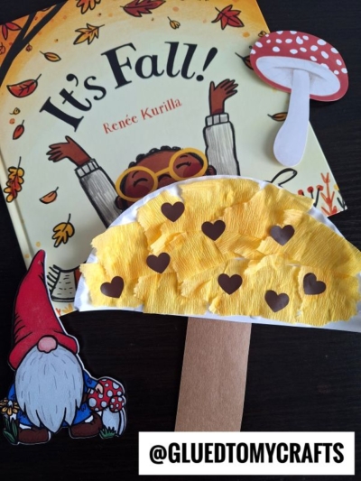 A fall-themed children’s book titled Its Fall! lies on a dark surface, surrounded by a gnome, a red mushroom, and charming paper plate mushrooms crafted with heart cutouts. Text: @GLUEDTOMYCRAFTS.