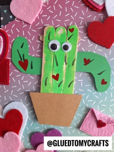 A handmade popsicle stick cactus craft for kids, featuring green-painted sticks, googly eyes, a brown paper pot, red hearts, and felt hearts on a pink and gray sprinkle-patterned background.