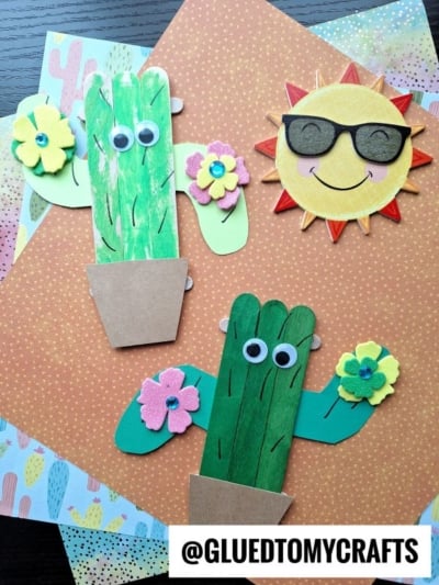 Handmade cactus craft for kids features green painted popsicle stick cactus with googly eyes in paper pots, topped with colorful paper flowers and placed beside a smiling sun craft. The background showcases patterned paper sheets.
