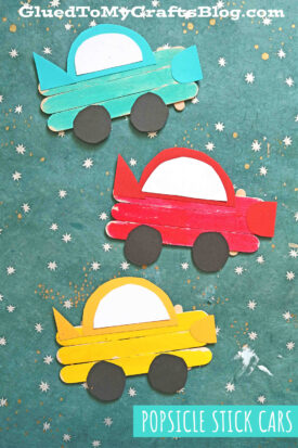 Three colorful craft stick cars made from popsicle sticks and paper sit on a green, star-patterned background. The cars are teal, red, and yellow with white windows and black wheels. A label reads “POPSICLE STICK CARS.”.
