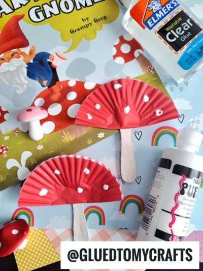 A craft project featuring cupcake liner mushrooms on sticks, white paint dots, glue bottles, a gnome-themed book, and a tag reading @GLUEDTOMYCRAFTS on a colorful background with rainbows—a whimsical mushroom craft for kids.
