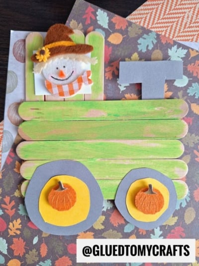A craft stick tractor made from green-painted popsicle sticks, grey paper wheels with orange pumpkin cutouts, and a smiling scarecrow figure, placed on autumn-themed paper backgrounds.
