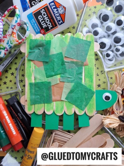 A Popsicle Stick Turtle craft made from green and tan tissue paper, colored craft sticks, and green paper with a googly eye on the head. Glue, scissors, markers, and other craft supplies surround it on a star-patterned surface.