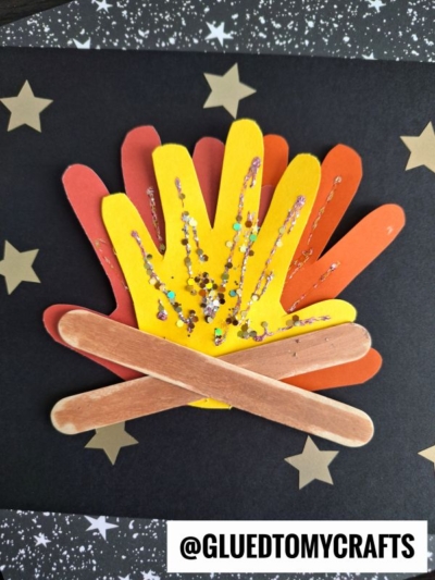 A Handprint Popsicle Stick Campfire made from red, orange, and yellow paper handprint flames, sparkling with glitter, sits atop crossed craft sticks as logs on a black background with gold stars.