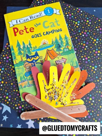 A children’s book titled Pete the Cat Goes Camping is on a star-patterned background. On top, there is a Handprint Popsicle Stick Campfire craft made from colored paper handprints and wooden sticks. @GLUEDTOMYCRAFTS is written at the bottom.