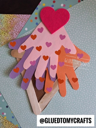 A layered paper craft features hand shapes in pink, peach, purple, and orange—like a sweet handprint ice cream treat—topped with a red heart and smaller heart stickers on colorful patterned paper. @GLUEDTOMYCRAFTS is written at the bottom.
