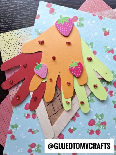 A colorful paper craft featuring cutouts of hand shapes in red, orange, yellow, and green—perfect for a fun handprint ice cream theme—decorated with strawberry stickers and glitter dots on floral paper. @GLUEDTOMYCRAFTS text is at the bottom.