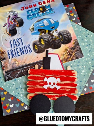 A children’s book titled “Elbow Grease: Fast Friends” sits on decorative paper. In front, a red popsicle stick craft shaped like a wagon—with black paper wheels and a white skull and crossbones—resembles popsicle stick monster trucks.