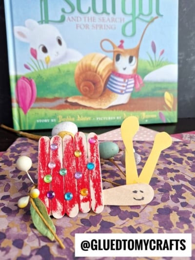 A snail kid craft made from red-painted popsicle stick snails, decorated with colorful gems, yellow paper antennae, and a smiling face sits on a flower-patterned surface, with a children’s book and small decorative items in the background.