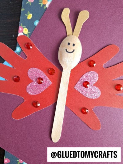 A charming spoon butterfly puppet made from a wooden spoon, red handprint cutouts as wings, pink glitter hearts, and red gems. This handprint wooden spoon butterfly is displayed on burgundy and floral paper with a smiling face drawn on the spoon.