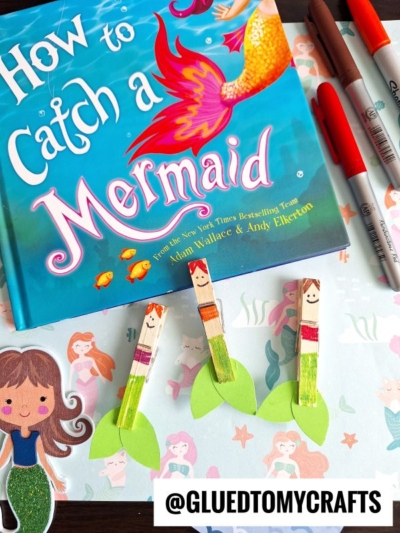 A colorful book titled How to Catch a Mermaid sits on a table with Sharpie markers and three Clothespin Mermaids, each with drawn faces and green paper tails, plus a mermaid sticker nearby.