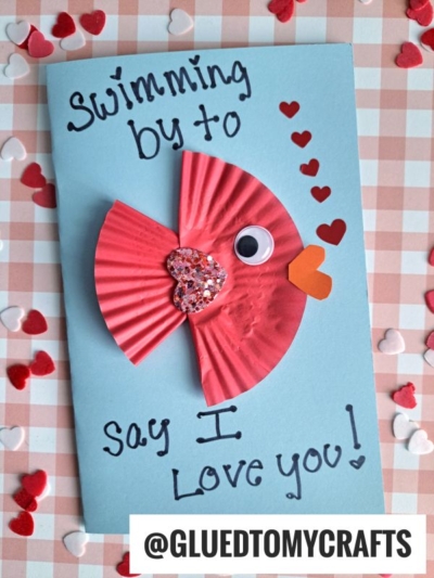 A blue cupcake liner fish card with a red cupcake liner fish, googly eye, and heart-shaped decorations. The text reads, Swimming by to say I love you! Red and white heart confetti are scattered on a checkered background.