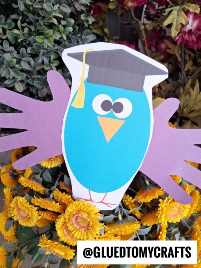 A blue paper graduation handprint owl with a graduation cap and purple handprint wings is displayed among yellow flowers and greenery. @GLUEDTOMYCRAFTS appears at the bottom.