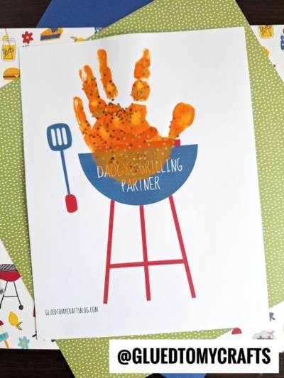 A children's craft featuring a handprint grill with an orange handprint as flames above a blue painted grill, labeled Handprint Daddy's Grilling Partner, displayed on patterned paper backgrounds.