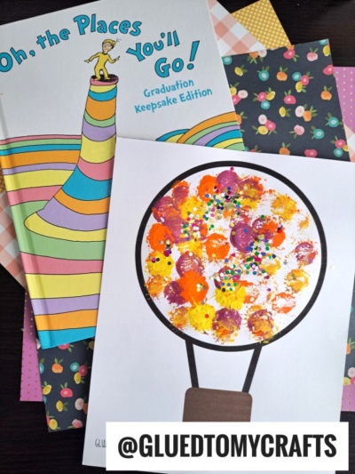 A colorful Dr. Seuss Oh, the Places You’ll Go! book sits next to a cheerful pom pom painted hot air balloon craft, displayed on patterned fruit paper. A tag reads @GLUEDTOMYCRAFTS.