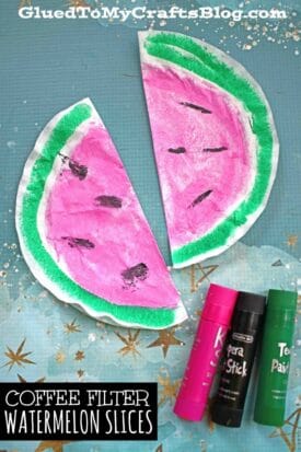 Two colorful coffee filter watermelon slices are displayed on a blue star-patterned background, with a glue stick and two paint bottles nearby. Text reads: COFFEE FILTER WATERMELON SLICES.