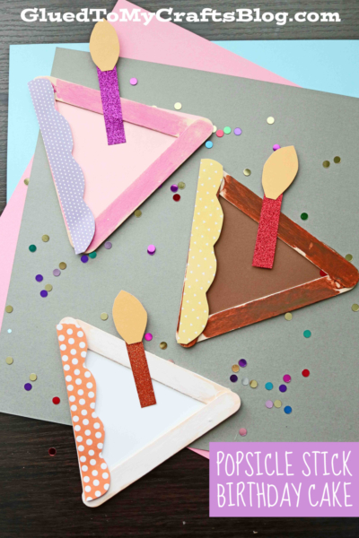Three triangular popsicle stick cake crafts, decorated with colored paper, glitter, and confetti, are arranged on pastel paper backgrounds. A text label reads Popsicle Stick Birthday Cake.