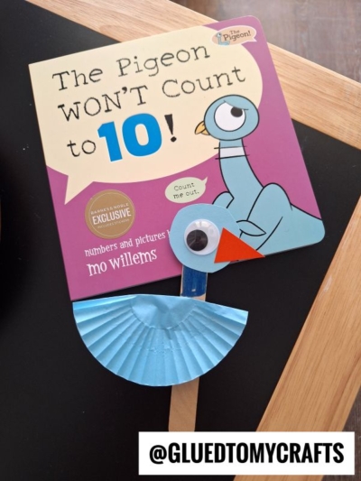 A children's book titled The Pigeon WONT Count to 10! by Mo Willems is decorated with a blue cupcake liner pigeon, complete with a googly eye and paper beak. @GLUEDTOMYCRAFTS is written at the bottom.