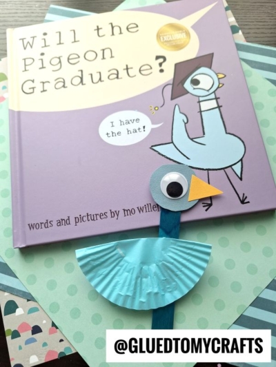 A children’s book titled “Will the Pigeon Graduate?” by Mo Willems sits under a handmade cupcake liner pigeon craft, made with a blue cupcake liner, googly eye, and craft stick. The handle @GLUEDTOMYCRAFTS appears at the bottom.