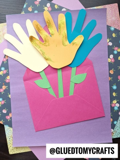 A handmade craft shows three handprint cards as flower tops in blue, yellow, and white, attached to green paper stems in a pink envelope on purple paper with floral sheets underneath. @GLUEDTOMYCRAFTS is written at the bottom.