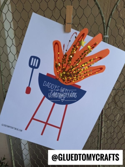 A printable craft shows a painted grill keepsake with red flames, gold glitter, and a star, labeled Daddy’s Grill. A spatula is next to the grill—perfect as a handprint daddy's grilling partner gift. @GLUEDTOMYCRAFTS username is at the bottom.