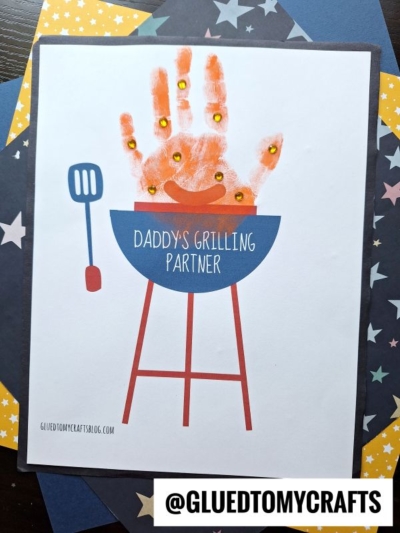 A handmade card features a blue handprint grill with red legs and a spatula, with an orange-painted handprint fire. Gold sequins decorate the flames. Text on the grill reads "Daddy's Grilling Partner." Star-patterned paper forms the background.