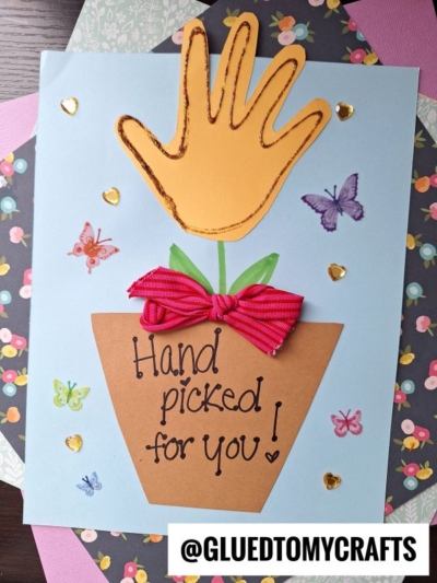 A handmade Handprint Flower Card features a flower pot with "Hand picked for you!" written on it, a yellow handprint as the flower, a red bow, butterflies, golden hearts, and the Instagram handle @GLUEDTOMYCRAFTS.