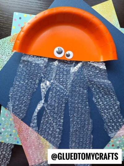 An orange paper plate with two googly eyes is decorated as a bubble wrap jelly fish, featuring bubble wrap tentacles hanging down. The jellyfish craft sits atop colorful, layered scrapbook paper.