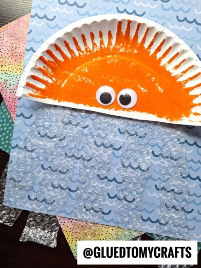 A kids’ craft features a paper plate painted orange like a sun with googly eyes, set on blue wave-patterned paper and partly covered with bubble wrap jelly fish to resemble water. Various colorful papers are in the background.