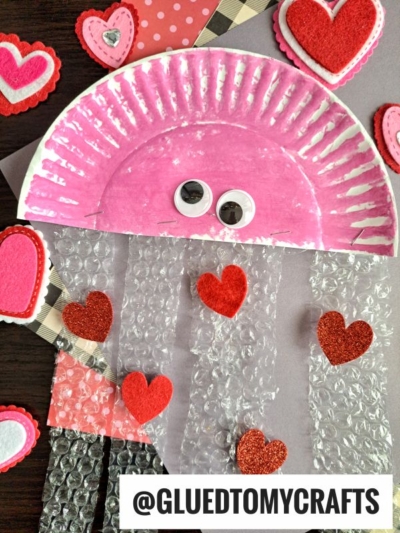 A pink bubble wrap jelly fish craft with googly eyes and strips of bubble wrap decorated with red glitter hearts, surrounded by felt heart shapes. The handle @GLUEDTOMYCRAFTS is visible at the bottom.