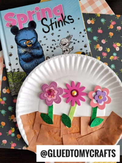 A paper plate spring scene featuring three pink foam flowers with green stems "planted" in brown paper dirt, set on floral paper beside the book Spring Stinks, starring a blue bear and white bunny. Text: @GLUEDTOMYCRAFTS.
