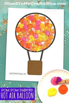A pom pom painted hot air balloon is decorated with colorful paint dots in orange, yellow, and pink made by dipping pom poms in paint. A small plate with paint and a pom pom appears in the lower right corner.