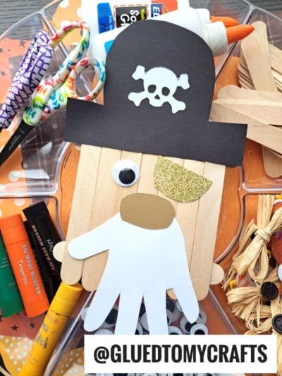 Create a Popsicle Stick Pirate using popsicle sticks, a white paper hand for a beard, a black pirate hat with skull and crossbones, googly eye, and gold glitter for the nose—surrounded by fun craft supplies!.